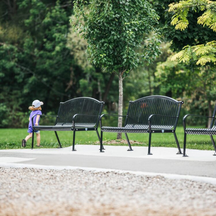a kid is happily running around the black classic steel garden and park benches in a park. The bench is ideal for commemorate a special event or a precious memory
